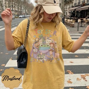 May include: A mustard yellow short-sleeve t-shirt featuring a colourful graphic print of Toy Story characters emerging from a purple bag. The design includes Woody, Buzz Lightyear, and other figures with the text Toy Story printed above the illustration.