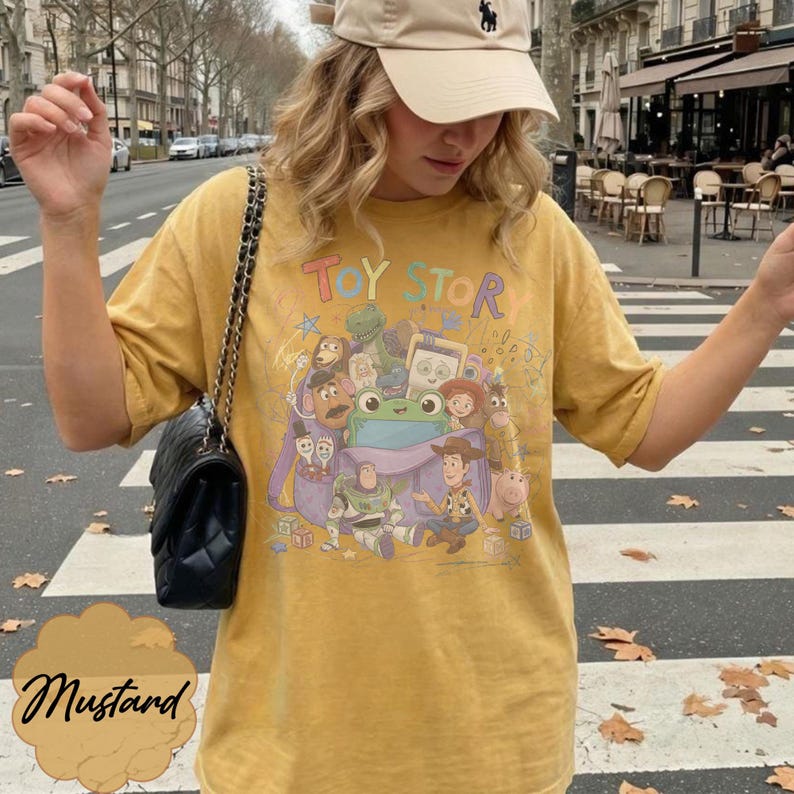 May include: A mustard yellow short-sleeve t-shirt featuring a colourful graphic print of Toy Story characters emerging from a purple bag. The design includes Woody, Buzz Lightyear, and other figures with the text Toy Story printed above the illustration.
