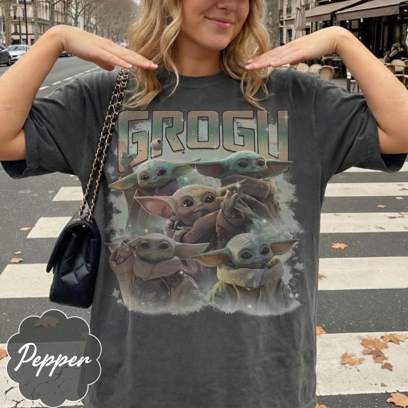 May include: A charcoal gray short-sleeve t-shirt featuring a graphic print of five small green creatures with large ears and eyes. The text GROGU is printed in a bold, distressed font above the characters, set against a soft, cloudy background.