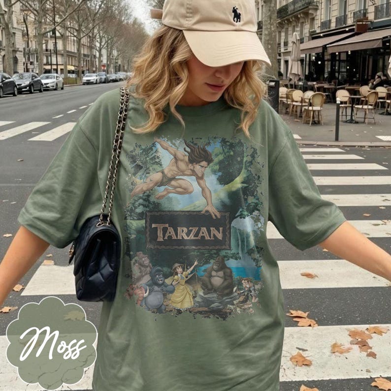 Op de afbeelding: Mosgroen T-shirt met een Tarzan-afbeelding. Het ontwerp bevat het woord "Tarzan" in een rechthoekige lijst, met illustraties van Tarzan en andere personages. Het shirt heeft een relaxte pasvorm.