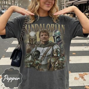 Pu&ograve; includere: Una t-shirt a maniche corte grigio antracite con una stampa grafica dei personaggi di The Mandalorian. Il design include il testo "MANDALORIAN" in lettere dorate sopra diverse figure corazzate e una piccola creatura verde con una tunica.