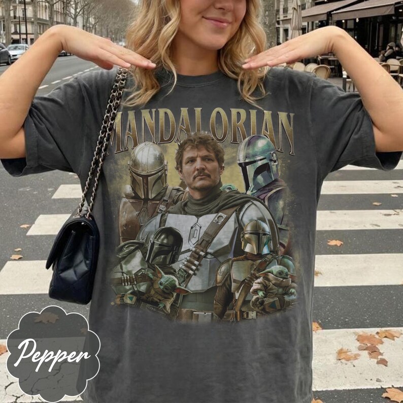 Pu&ograve; includere: Una t-shirt a maniche corte grigio antracite con una stampa grafica dei personaggi di The Mandalorian. Il design include il testo "MANDALORIAN" in lettere dorate sopra diverse figure corazzate e una piccola creatura verde con una tunica.