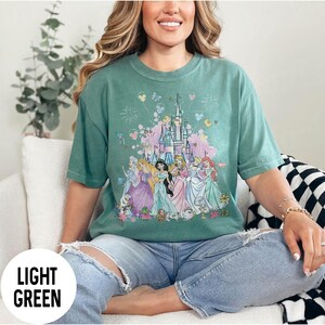 May include: Light green t-shirt featuring a graphic of a castle and several princesses. The shirt has a crew neck and short sleeves. The words "LIGHT GREEN" are printed on a white circle in the lower left corner.
