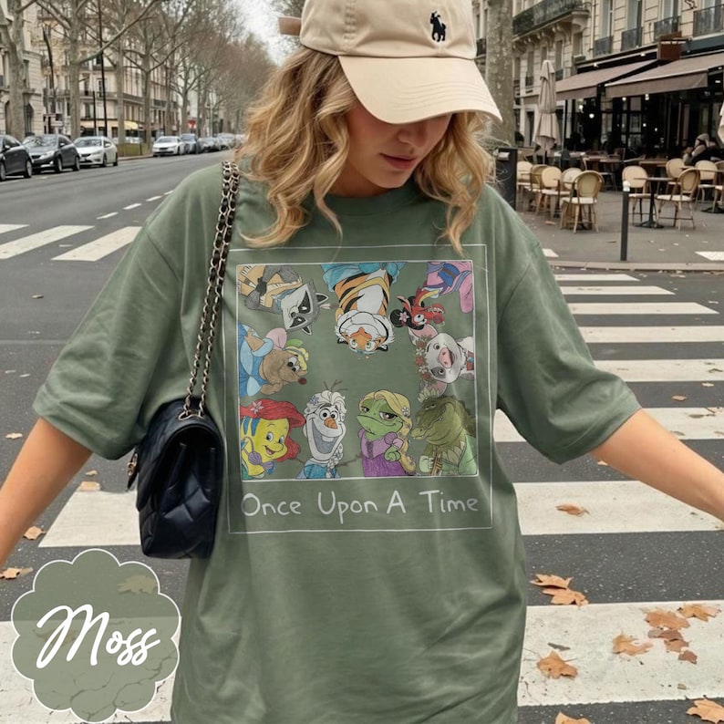 May include: Moss green t-shirt featuring a colorful graphic of cartoon characters with the text "Once Upon A Time". The shirt has short sleeves and a relaxed fit. A black purse with a chain strap is visible.