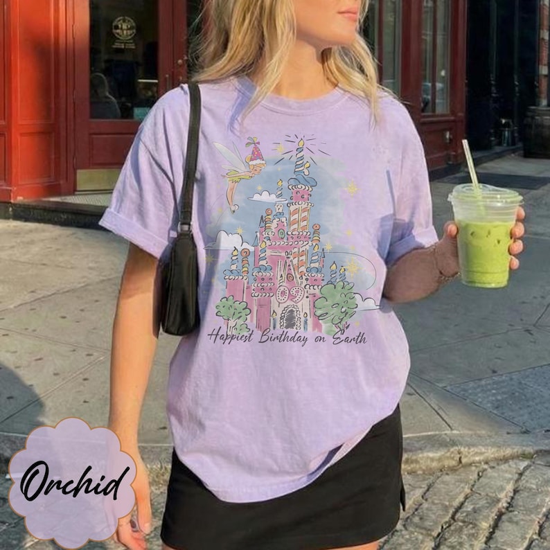 Puede incluir: Una camiseta de color lila claro con un gr&aacute;fico de un castillo de cuento de hadas, un hada y el texto "Happiest Birthday on Earth". La persona lleva una camiseta y sostiene una bebida verde en un vaso de pl&aacute;stico transparente.