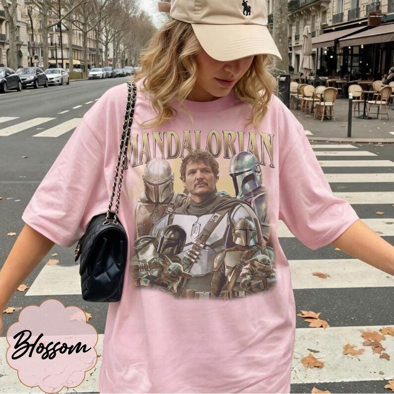 Pu&ograve; includere: Una t-shirt rosa chiaro con una stampa grafica dei personaggi di The Mandalorian, tra cui la figura centrale, guerrieri in armatura e una piccola creatura verde. Il testo "MANDALORIAN" &egrave; stampato in un carattere dorato sopra l'illustrazione.