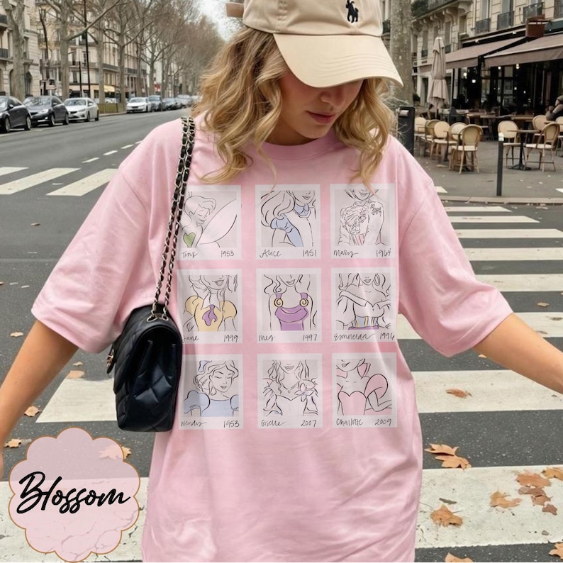 May include: A light pink t-shirt featuring a grid of cartoon illustrations of Disney princesses with their names and years. The shirt has short sleeves and a relaxed fit. The word "Blossom" is printed on the bottom left.