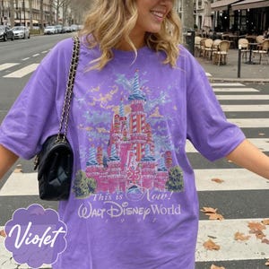 K&ouml;nnte beinhalten: Ein violettes Kurzarm-T-Shirt mit einer Grafik eines rosafarbenen Schlosses mit rot-wei&szlig; gestreiften T&uuml;rmen. Der Text lautet "25", "This is Now!", "Walt Disney World" und "1997". Eine schwarze gesteppte Handtasche mit goldener Kette ist zu sehen.