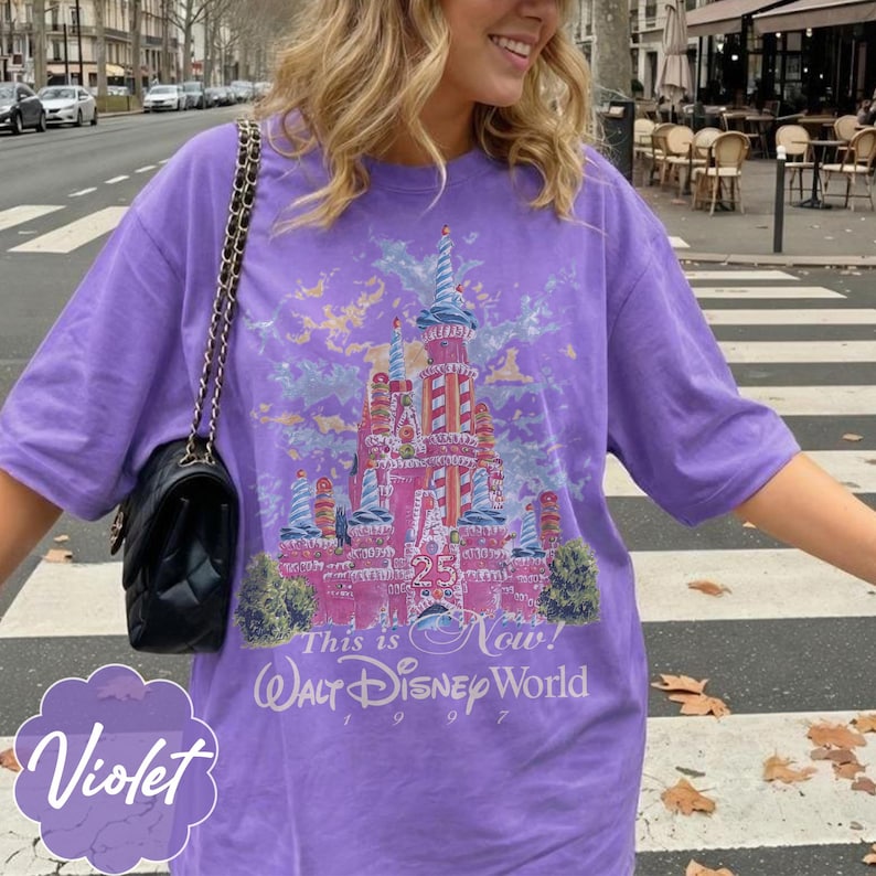 K&ouml;nnte beinhalten: Ein violettes Kurzarm-T-Shirt mit einer Grafik eines rosafarbenen Schlosses mit rot-wei&szlig; gestreiften T&uuml;rmen. Der Text lautet "25", "This is Now!", "Walt Disney World" und "1997". Eine schwarze gesteppte Handtasche mit goldener Kette ist zu sehen.