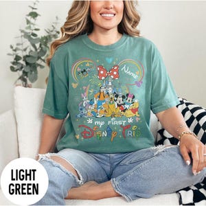 May include: Light green t-shirt with a colorful graphic featuring Disney characters, a castle, and the text "My First Disney Trip." The design includes a large red bow and the word "Name."
