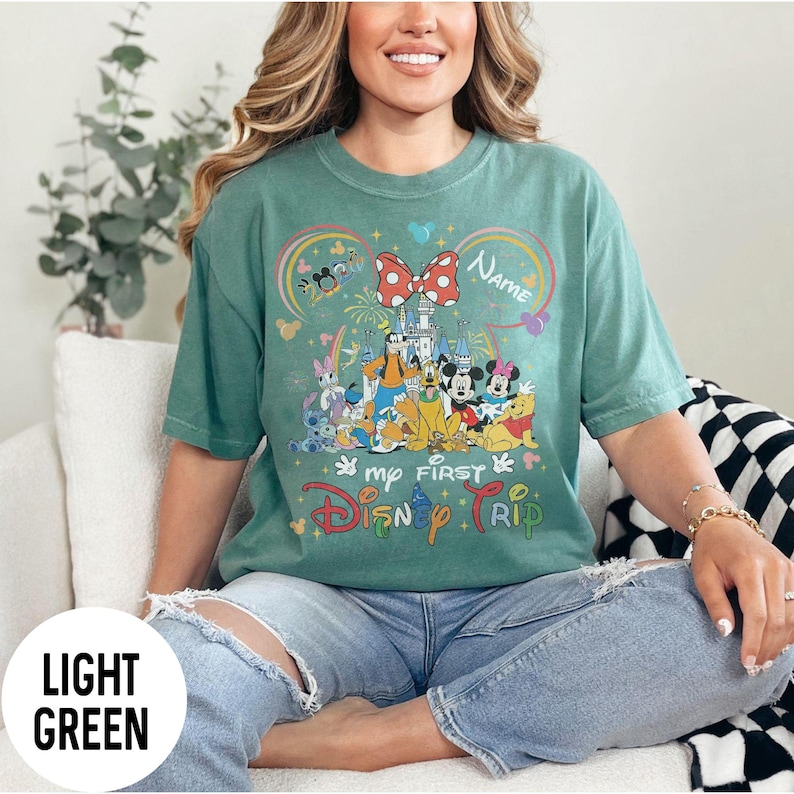 May include: Light green t-shirt with a colorful graphic featuring Disney characters, a castle, and the text "My First Disney Trip." The design includes a large red bow and the word "Name."