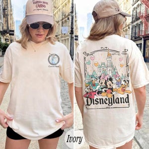Pu&ograve; includere: T-shirt color avorio con grafica anteriore e posteriore. La grafica anteriore presenta un'immagine di cartone animato all'interno di un cerchio a scacchi. La grafica posteriore mostra un design di Disneyland con personaggi dei cartoni animati e la scritta "Disneyland 1955".