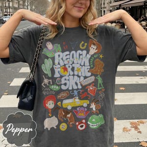 Pode incluir: Camiseta cinza escuro com um gr&aacute;fico colorido com personagens e texto do filme "Toy Story". O design inclui a frase "Reach for the Sky" e v&aacute;rias ilustra&ccedil;&otilde;es de desenhos animados.