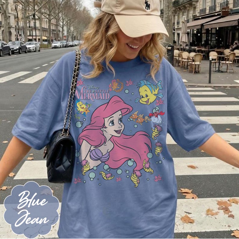 May include: Blue jean-coloured t-shirt featuring a graphic of Ariel from Disney's The Little Mermaid. The design includes Ariel with pink hair, a purple seashell bra, and various sea creatures. The shirt has short sleeves.