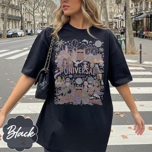 May include: Black t-shirt with a vibrant graphic depicting Universal Studios attractions and characters. The text "UNIVERSAL STUDIOS" is displayed prominently. The word "Black" is written in a decorative font on the bottom left of the shirt.