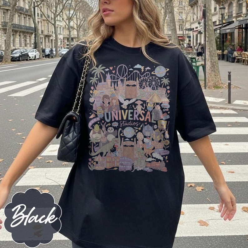 May include: Black t-shirt with a vibrant graphic depicting Universal Studios attractions and characters. The text "UNIVERSAL STUDIOS" is displayed prominently. The word "Black" is written in a decorative font on the bottom left of the shirt.