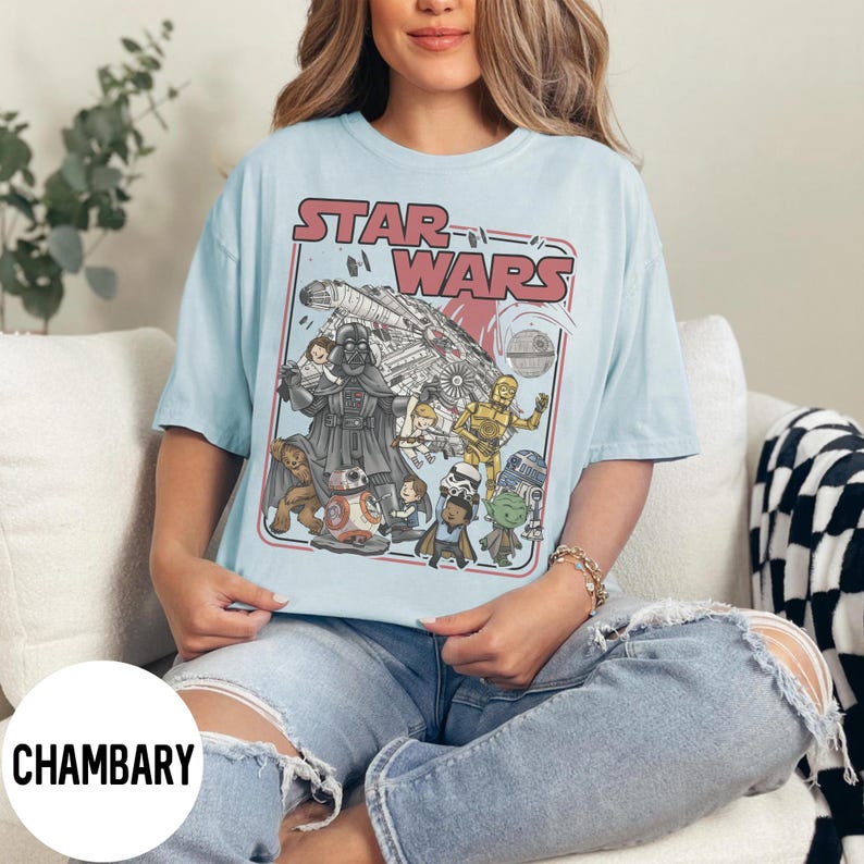 May include: Light blue Star Wars graphic t-shirt featuring Darth Vader, C-3PO, R2-D2, and other characters. The shirt has the words "STAR WARS" in a red retro font. The shirt is a short-sleeved, crew neck style.