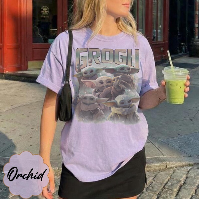 May include: A light purple t-shirt featuring a graphic print of multiple Grogu characters and the text GROGU in a bold font. The shirt has short sleeves and a crew neckline, paired with a black shoulder bag and a clear plastic cup of green drink.