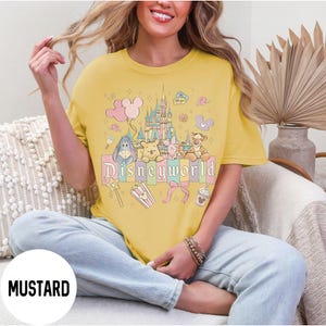 May include: Mustard yellow t-shirt with a colorful Disneyworld graphic. The graphic features a castle, balloons, and cartoon characters including Winnie the Pooh, Eeyore, Piglet, and Tigger. The word "Disneyworld" is printed in a pastel font.