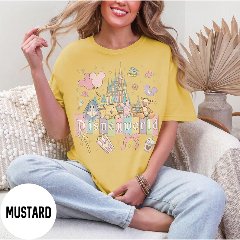 May include: Mustard yellow t-shirt with a colorful Disneyworld graphic. The graphic features a castle, balloons, and cartoon characters including Winnie the Pooh, Eeyore, Piglet, and Tigger. The word "Disneyworld" is printed in a pastel font.