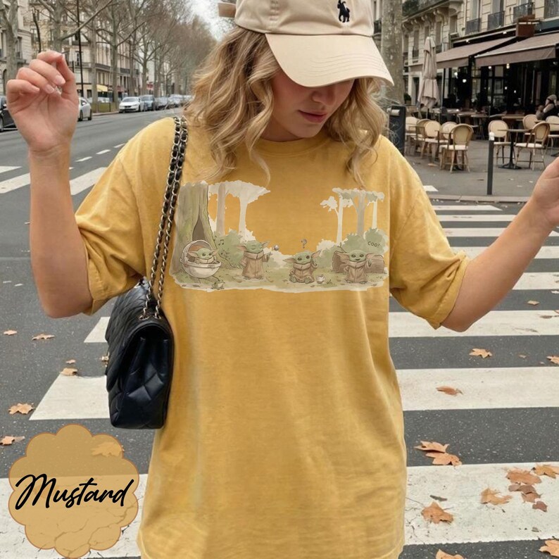 Puede incluir: Camiseta de color mostaza con un dise&ntilde;o de Grogu de The Mandalorian. El dise&ntilde;o muestra a Grogu con otros personajes en un entorno forestal. La camiseta tiene mangas cortas y un corte holgado. La palabra "Mustard" est&aacute; escrita en cursiva en la parte inferior.