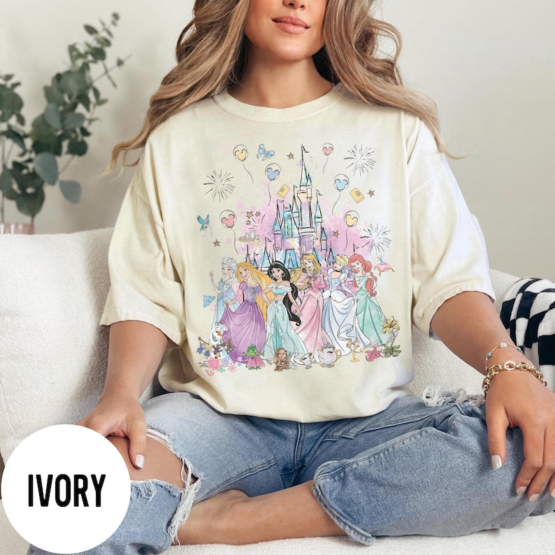 May include: Ivory-colored t-shirt featuring a colorful illustration of Disney princesses in front of a castle. The design includes fireworks, balloons, and floral accents. The word "IVORY" is printed on a white circle.