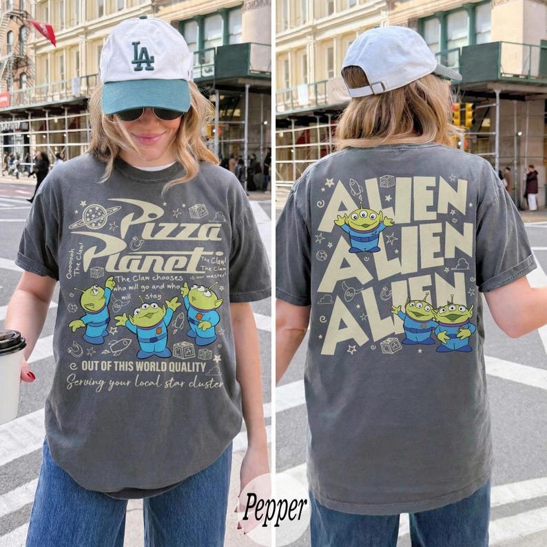 K&ouml;nnte beinhalten: Dunkelgraues T-Shirt mit dem Pizza Planet-Logo und drei gr&uuml;nen Aliens. Auf der Vorderseite des Shirts steht "Pizza Planet" und "Out of this world quality". Auf der R&uuml;ckseite des Shirts steht das Wort "ALIEN" mehrfach.