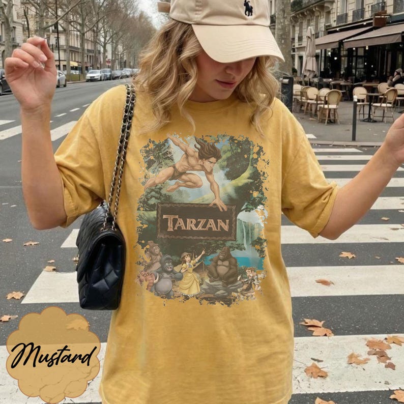 Op de afbeelding: Mosterdkleurig T-shirt met een Tarzan-afbeelding. Het ontwerp toont Tarzan, junglesc&egrave;nes en het woord "Tarzan" op een houten bord. Het shirt is een casual stijl met korte mouwen.