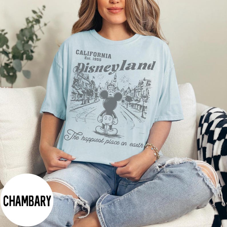 Może przedstawiać: Jasnoniebieska koszulka z grafiką Disneyland. Grafika przedstawia Myszka Miki przed sceną uliczną z napisem "CALIFORNIA Est. 1955 Disneyland" i "The happiest place on earth."