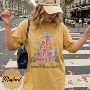 K&ouml;nnte beinhalten: Ein senfgelbes Kurzarm-T-Shirt mit einem Grafikdruck eines rosa Schlosses und dem Text "This is the Magic!" sowie "Walt Disney World" darunter. Das Shirt hat einen Rundhalsausschnitt und eine lockere Passform, kombiniert mit einer schwarzen gesteppten Umh&auml;ngetasche.
