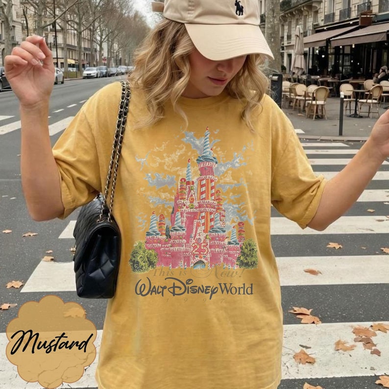 K&ouml;nnte beinhalten: Ein senfgelbes Kurzarm-T-Shirt mit einem Grafikdruck eines rosa Schlosses und dem Text "This is the Magic!" sowie "Walt Disney World" darunter. Das Shirt hat einen Rundhalsausschnitt und eine lockere Passform, kombiniert mit einer schwarzen gesteppten Umh&auml;ngetasche.