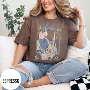 Może przedstawiać: Brązowa koszulka w kolorze espresso z grafiką zamku, balon&oacute;w Myszki Miki i niebieskiej kokardy, obramowanej kwiatowymi akcentami. Koszulka ma luźny kr&oacute;j i kr&oacute;tkie rękawy. Słowo "ESPRESSO" jest wydrukowane na dole po lewej stronie.