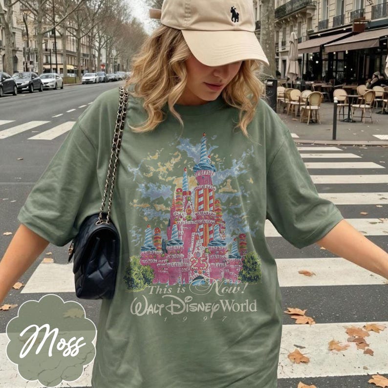 K&ouml;nnte beinhalten: Ein olivgr&uuml;nes T-Shirt mit einer Grafik eines rosafarbenen Schlosses mit blauen Akzenten sowie dem Text "25", "This is Now!", "WALT DISNEY WORLD" und "1997". Eine beige Baseballkappe mit kleinem dunklen Logo und eine schwarze gesteppte Umh&auml;ngetasche.
