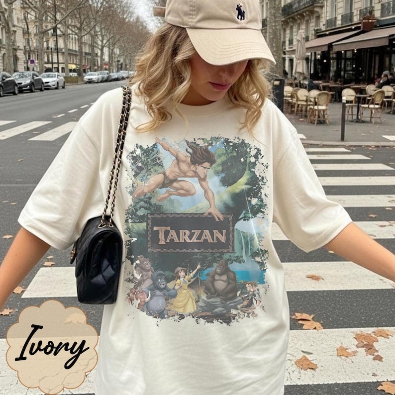 Op de afbeelding: Ivoor oversized T-shirt met een Tarzan-afbeelding. De afbeelding bevat het woord "Tarzan" in een rechthoekig kader, met een afbeelding van Tarzan en andere personages. Het shirt heeft korte mouwen.
