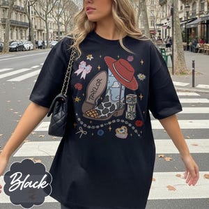 May include: A black short-sleeve t-shirt featuring a graphic print of a cowboy boot with a cow print pattern, a red hat, and blocks with the letters T, S, and the number 13. The word TAYLOR is printed on the boot, surrounded by small stars and bows.