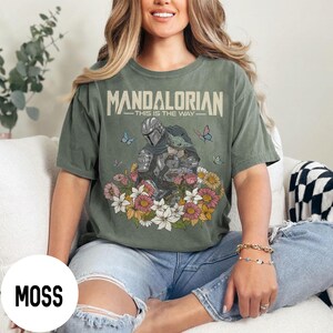 Puede incluir: Camiseta verde musgo con un dise&ntilde;o de The Mandalorian y Grogu, rodeados de flores y mariposas. El texto "MANDALORIAN - THIS IS THE WAY" est&aacute; impreso en la parte superior. La camiseta es de corte holgado.