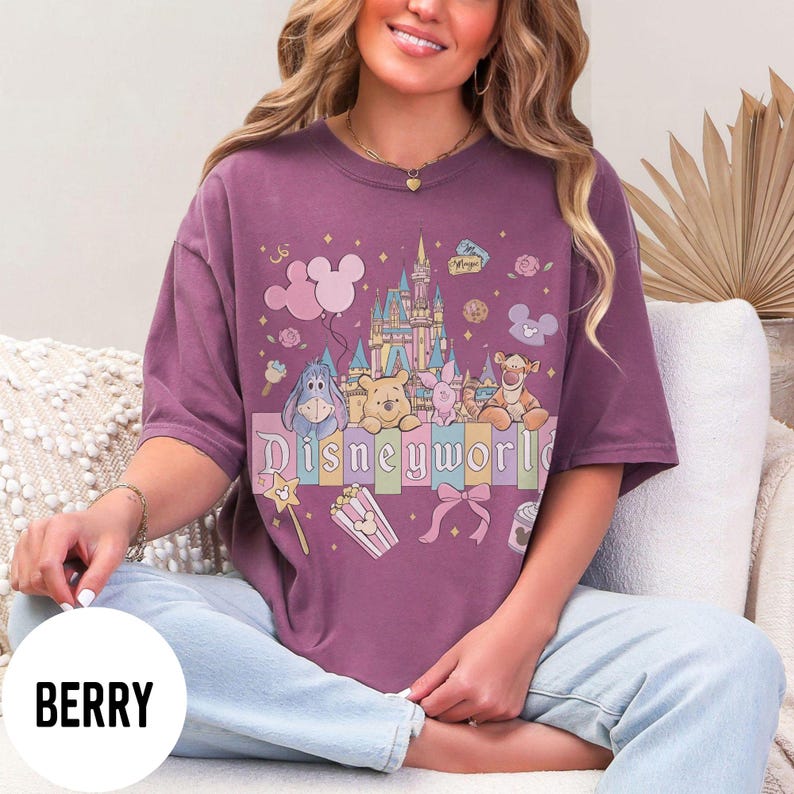 May include: Berry-colored t-shirt featuring a colorful Disney World graphic. The design includes a castle, balloons, and various Disney characters like Winnie the Pooh and Eeyore. The word "Disneyworld" is displayed in pastel colors.