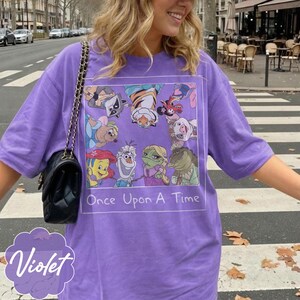 May include: Violet-colored t-shirt with a graphic featuring cartoon characters and the text "Once Upon A Time." The shirt has short sleeves and a relaxed fit. A black purse with a chain strap is visible.