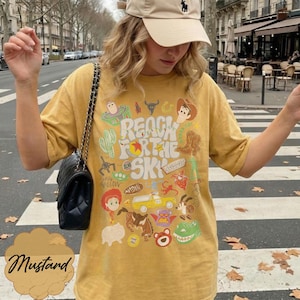 Pode incluir: Camiseta mostarda com um design gr&aacute;fico com personagens de desenhos animados e o texto "REACH FOR THE SKY". A palavra "Mustard" est&aacute; escrita em it&aacute;lico no canto inferior esquerdo.