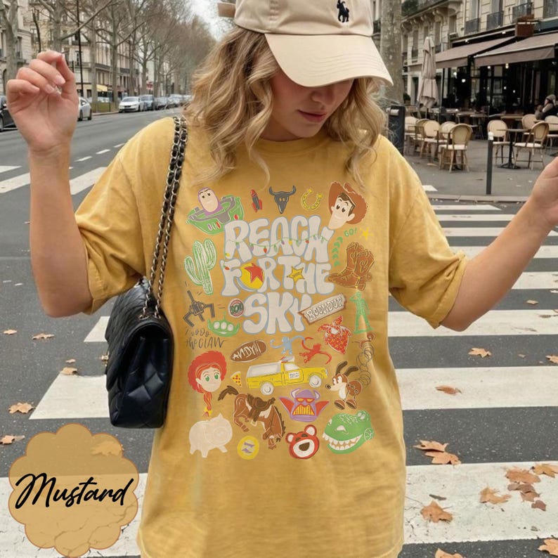 Pode incluir: Camiseta mostarda com um design gr&aacute;fico com personagens de desenhos animados e o texto "REACH FOR THE SKY". A palavra "Mustard" est&aacute; escrita em it&aacute;lico no canto inferior esquerdo.
