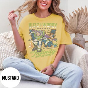 May include: Mustard yellow t-shirt with a graphic of Buzz Lightyear and Woody from Toy Story. The design includes the text "Buzz & Woody Lightyear" and "To Infinity and Beyond". The shirt has a relaxed fit.