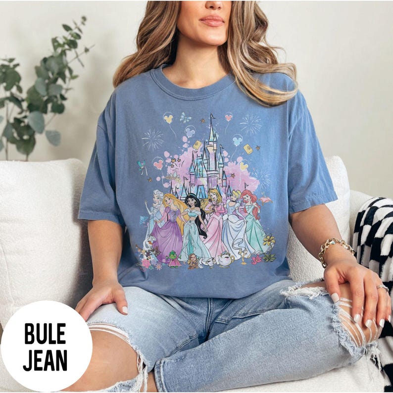 May include: A blue t-shirt featuring a colorful graphic of a castle and several princesses. The shirt has short sleeves and a relaxed fit. The text "BULE JEAN" is in a white circle on the bottom left.