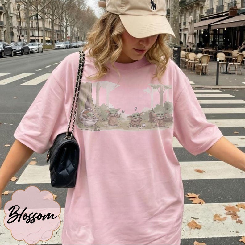 Puede incluir: Camiseta rosa claro con un gr&aacute;fico de Grogu de The Mandalorian. El dise&ntilde;o muestra a Grogu en una cesta y con otros personajes. La palabra "Blossom" est&aacute; en cursiva en la parte inferior izquierda.