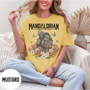 Puede incluir: Camiseta de color mostaza con el texto "MANDALORIAN - THIS IS THE WAY" sobre una ilustraci&oacute;n del Mandaloriano y Grogu, rodeados de flores y mariposas. La camiseta es de corte holgado.