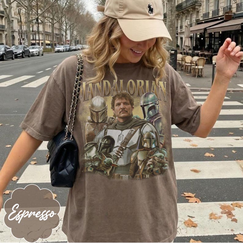 Pu&ograve; includere: Una t-shirt marrone a maniche corte con una stampa grafica dei personaggi di The Mandalorian, inclusa una figura centrale e diversi elmi. La maglia &egrave; abbinata a una borsa a tracolla nera trapuntata con una catena color argento.