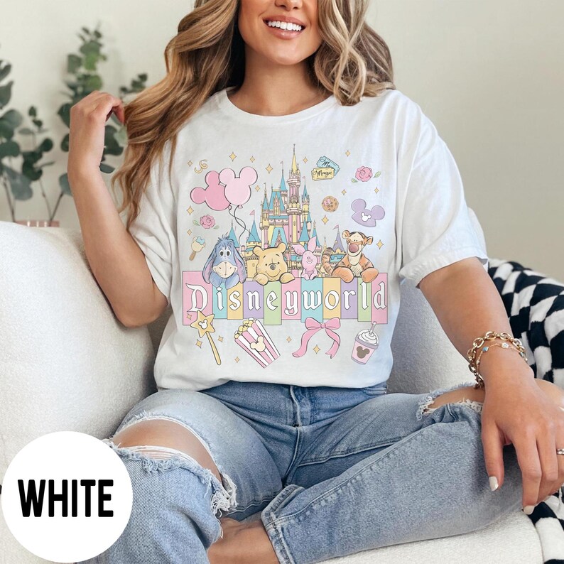 May include: White t-shirt featuring a colorful Disneyworld graphic. The design includes a castle, cartoon characters, and the word "Disneyworld" in pastel colors. The shirt is a relaxed fit.