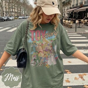 May include: An olive green t-shirt featuring a graphic print of various animated characters and the text "TOY story" in pink and blue lettering. The shirt has short sleeves and a crew neckline, paired with a black quilted shoulder bag with a chain strap.