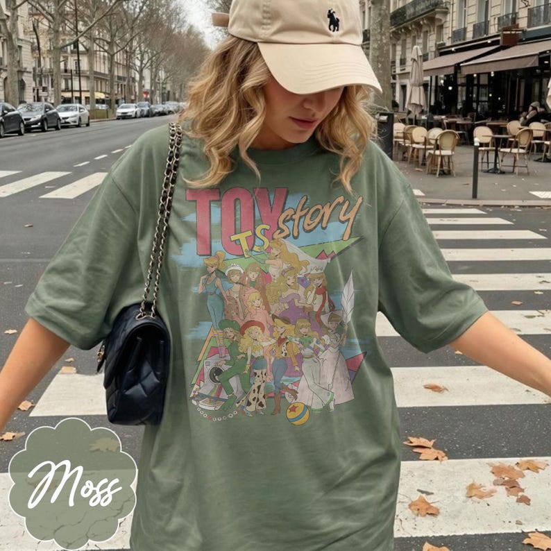 May include: An olive green t-shirt featuring a graphic print of various animated characters and the text "TOY story" in pink and blue lettering. The shirt has short sleeves and a crew neckline, paired with a black quilted shoulder bag with a chain strap.