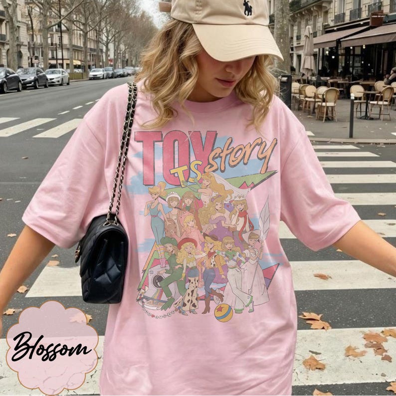 May include: A light pink short-sleeved t-shirt featuring a graphic print of various female characters from Toy Story with the text Toy Story in large letters. The shirt is paired with a black quilted shoulder bag with a silver chain strap.