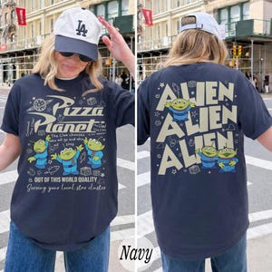 K&ouml;nnte beinhalten: Marineblaues T-Shirt mit einem Pizza Planet-Motiv auf der Vorderseite und Alien-Charakteren auf der R&uuml;ckseite. Das Frontdesign enth&auml;lt den Text "Pizza Planet" und Cartoon-Aliens. Das R&uuml;ckendesign zeigt das Wort "Alien" dreimal wiederholt.